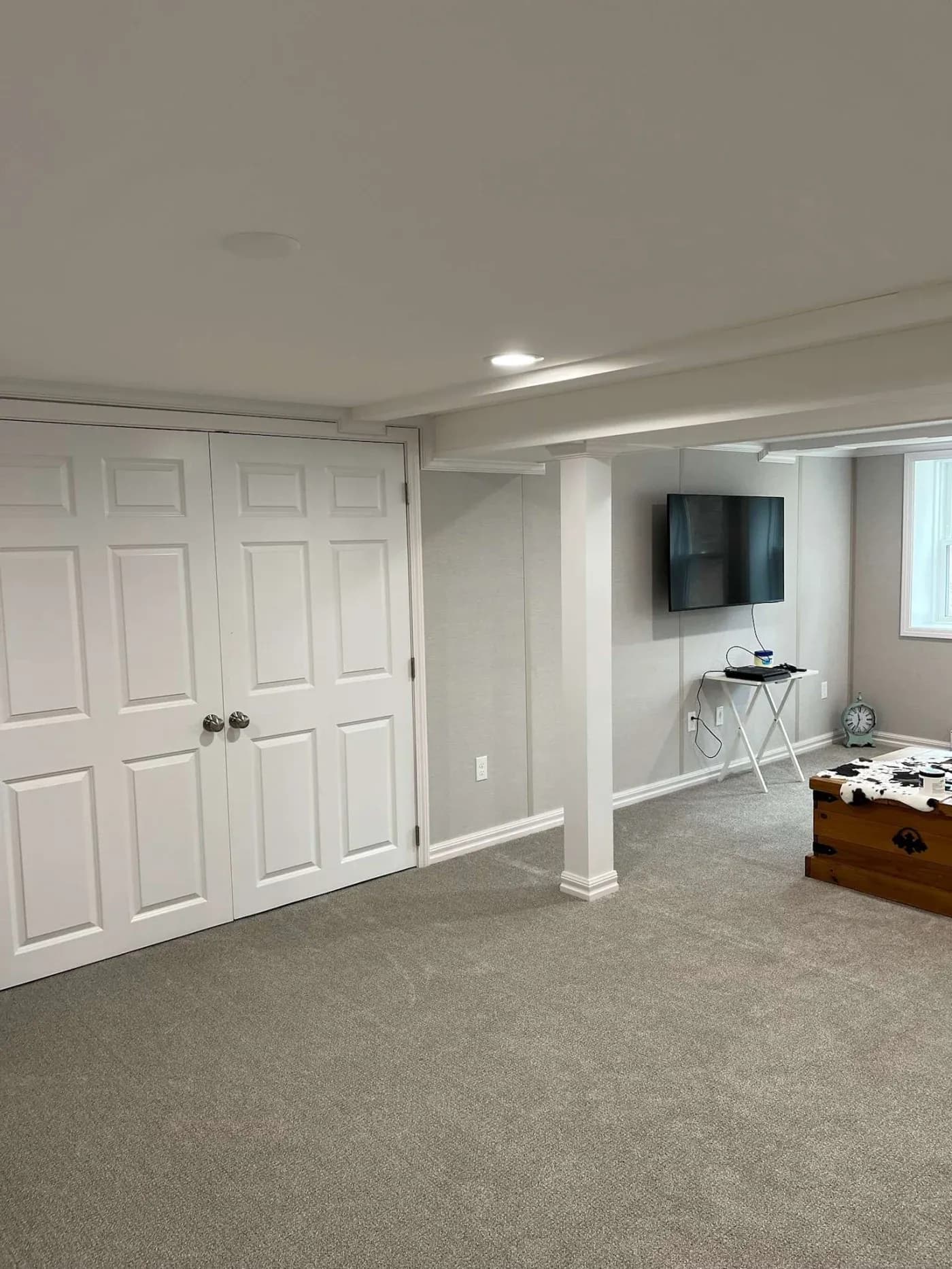 A cozy basement room with beige carpet, white double doors, a wall-mounted TV, and a small side table. Natural light filters through a window, creating a tranquil atmosphere.