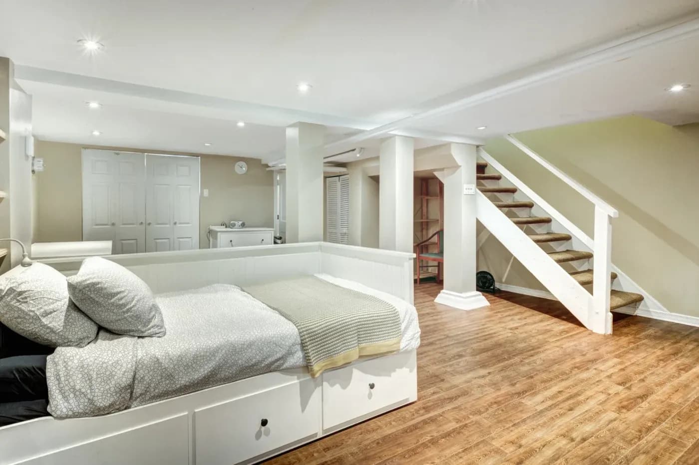 A bright, modern basement bedroom with a white daybed, soft pillows, and a knitted throw. Wooden floors and a staircase enhance the cozy, welcoming feel.