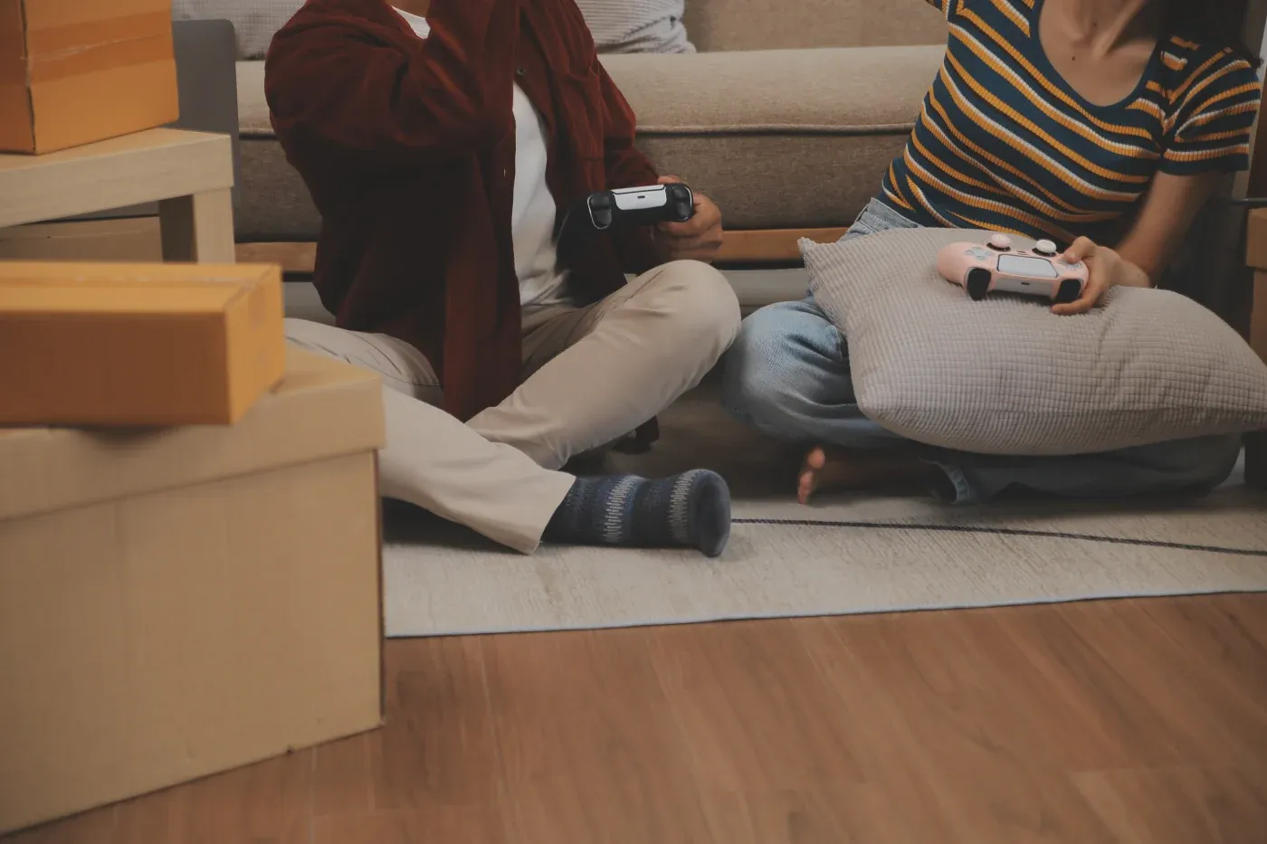 Two people sit on the floor by a couch, holding game controllers. They're surrounded by cardboard boxes, suggesting a moving day. Cozy and relaxed mood.