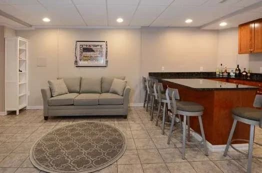 Spacious basement with a gray sofa, round patterned rug, fully stocked bar with high stools, and warm lighting, creating a cozy, inviting atmosphere.