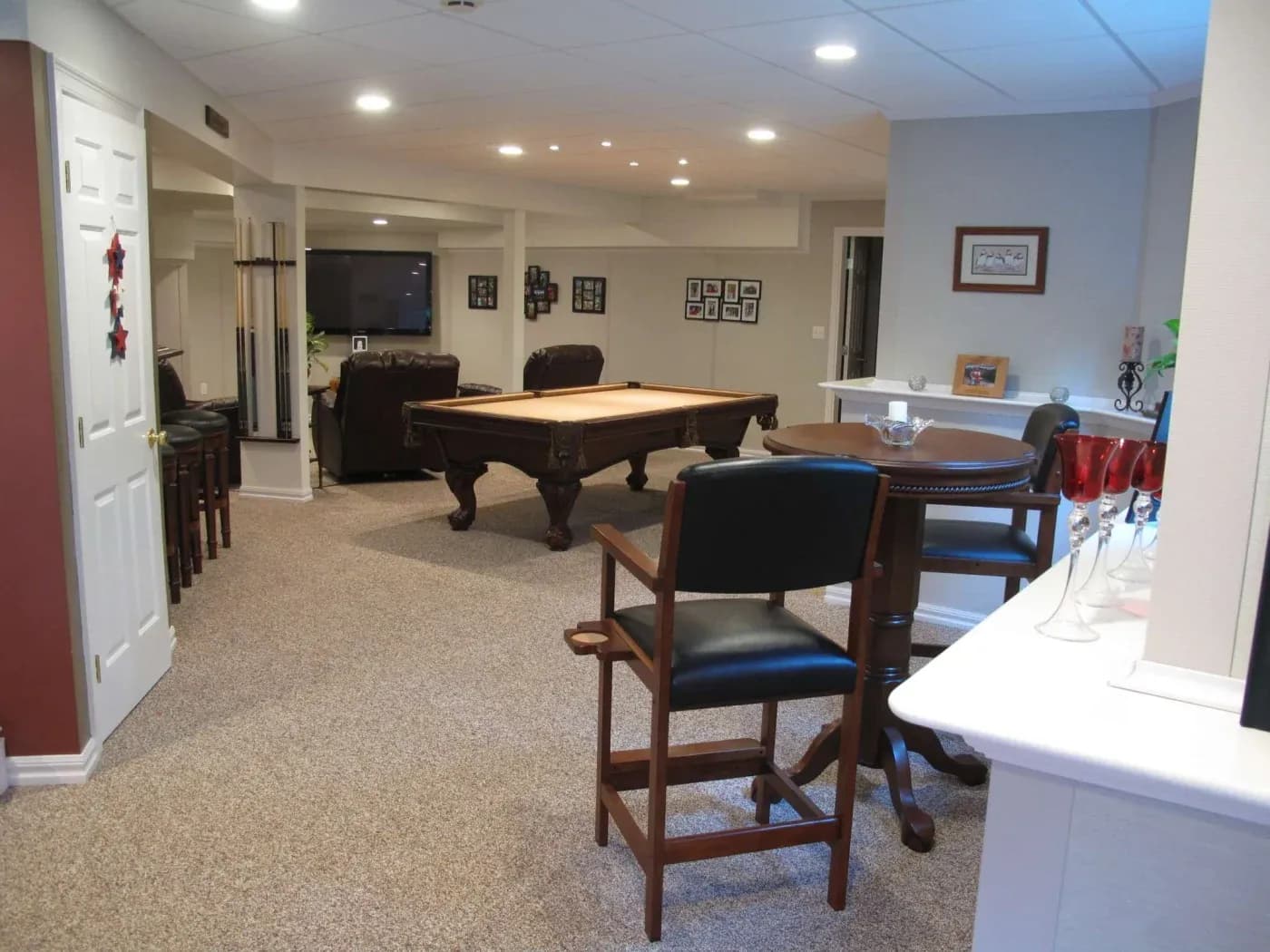 Spacious basement game room with a pool table, bar stools, and a TV on the wall. Cozy lighting and framed photos create a welcoming atmosphere.