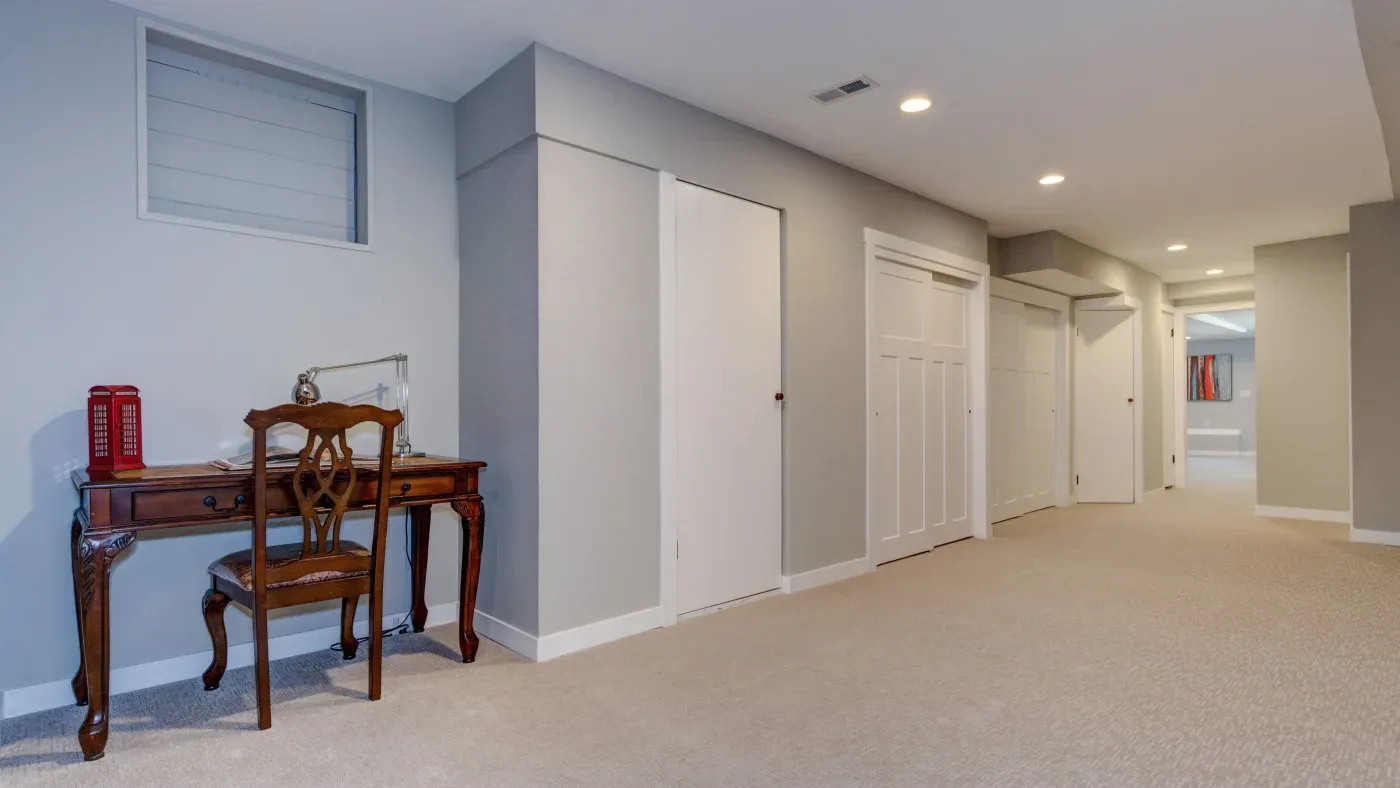 Spacious modern hallway with beige carpet, gray walls, and recessed lighting. A wooden desk with a chair is on the left, creating a cozy workspace.
