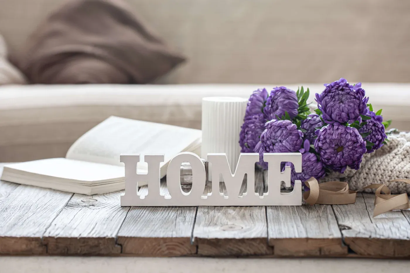Decorative "HOME" letters on a rustic wooden table, surrounded by an open book, a candle, and a bouquet of purple flowers, creating a cozy atmosphere.