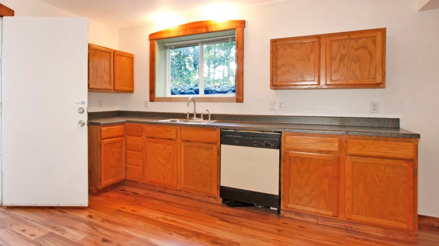 A kitchen with wooden cabinets, beige countertop, and a window above a dual sink. A dishwasher is to the right, with warm lighting and wooden flooring.
