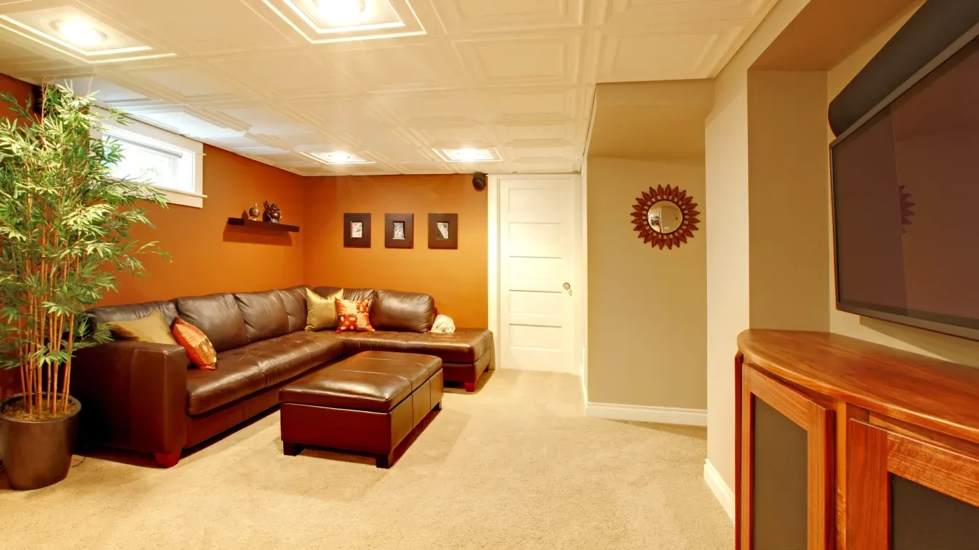 Cozy basement with a white ceiling and soft lighting. Features a brown leather sectional, potted plant, and wall decor, creating a warm, inviting ambiance.