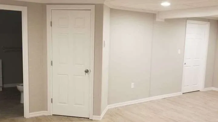 A basement with light beige walls and wooden flooring, featuring two white doors. The room appears minimalistic and clean, with a toilet visible in a doorway.