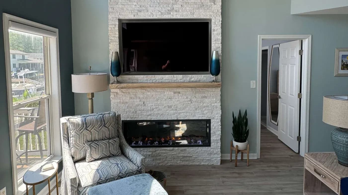 A cozy living room features a stone fireplace with a sleek TV above. A patterned armchair and lamp sit by a large window, creating a bright, inviting space.