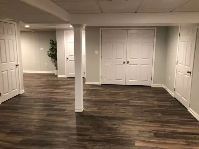 Spacious room with wooden floors and white paneled walls. Several closed white doors are visible, with two support columns in the center and a potted plant in the corner.