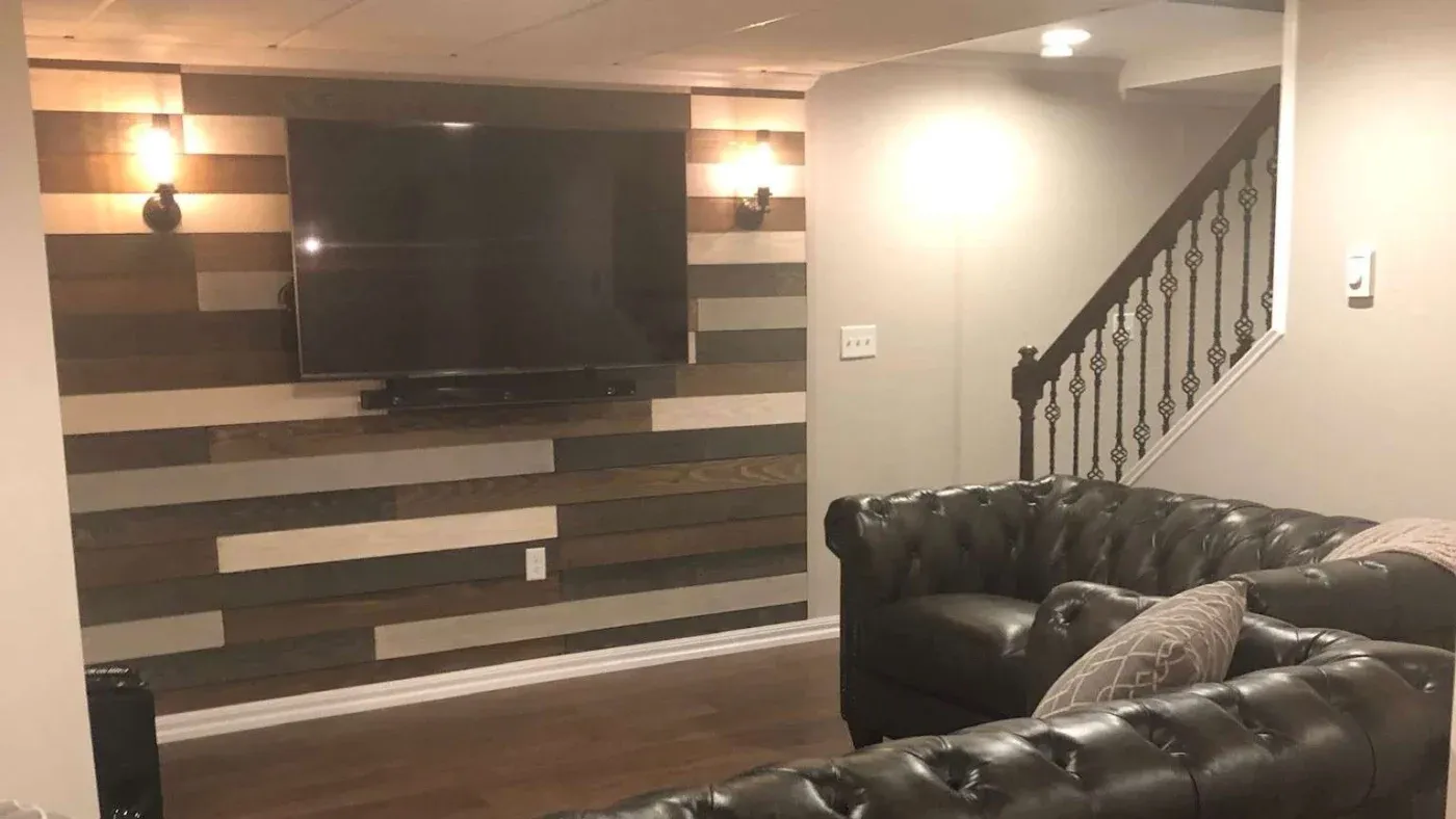 Cozy basement living room with tufted leather sofas, a large flat-screen TV on a striped wood accent wall, warm lighting, and a staircase with ornate railing.