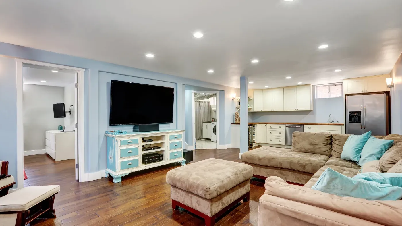 A spacious, cozy living area with light blue walls, a large beige sectional sofa, and teal cushions. A large TV is mounted above a vintage turquoise console. The kitchen, visible in the background, features modern appliances and white cabinetry. The room's warm wooden flooring adds to the inviting atmosphere.
