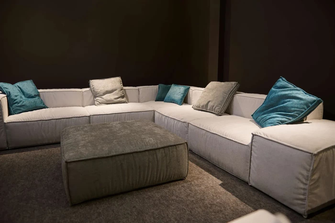 Spacious L-shaped sofa with soft, gray fabric and teal accent pillows, placed in a cozy, dimly lit room. A matching square ottoman is centered in front.