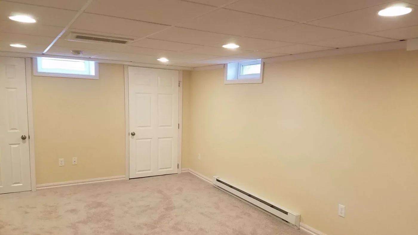 A clean, empty basement room with beige walls, white ceiling tiles, and soft carpet. Two small windows and a closed white door are visible, creating a cozy atmosphere.