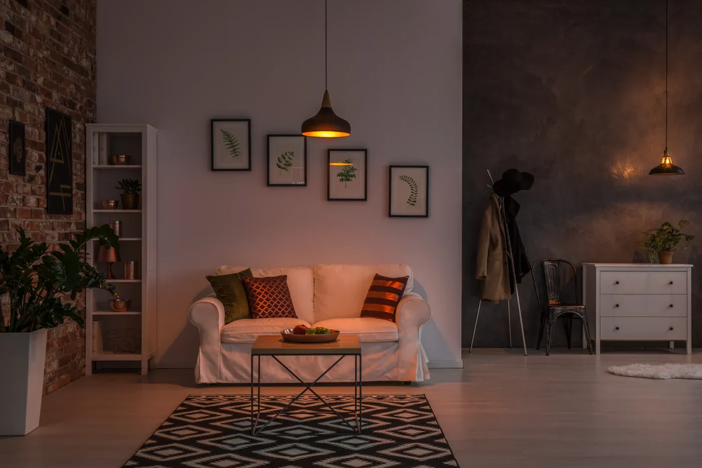 Cozy living room with a white sofa, colorful pillows, and a coffee table under warm pendant light. Framed leaf art decorates the wall.