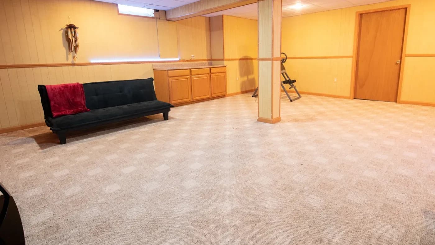 A spacious basement with beige carpet and wood-paneled walls features a dark futon with a red blanket, wooden cabinets, and a door. It has a calm, minimalist feel.