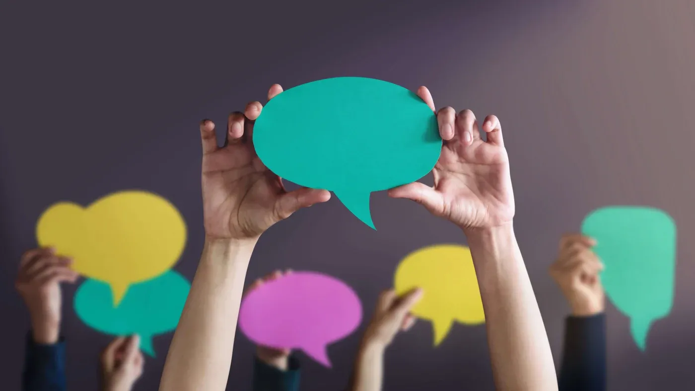 Hands hold colorful paper speech bubbles against a dark background, symbolizing communication and diverse conversation. The tone is lively and interactive.