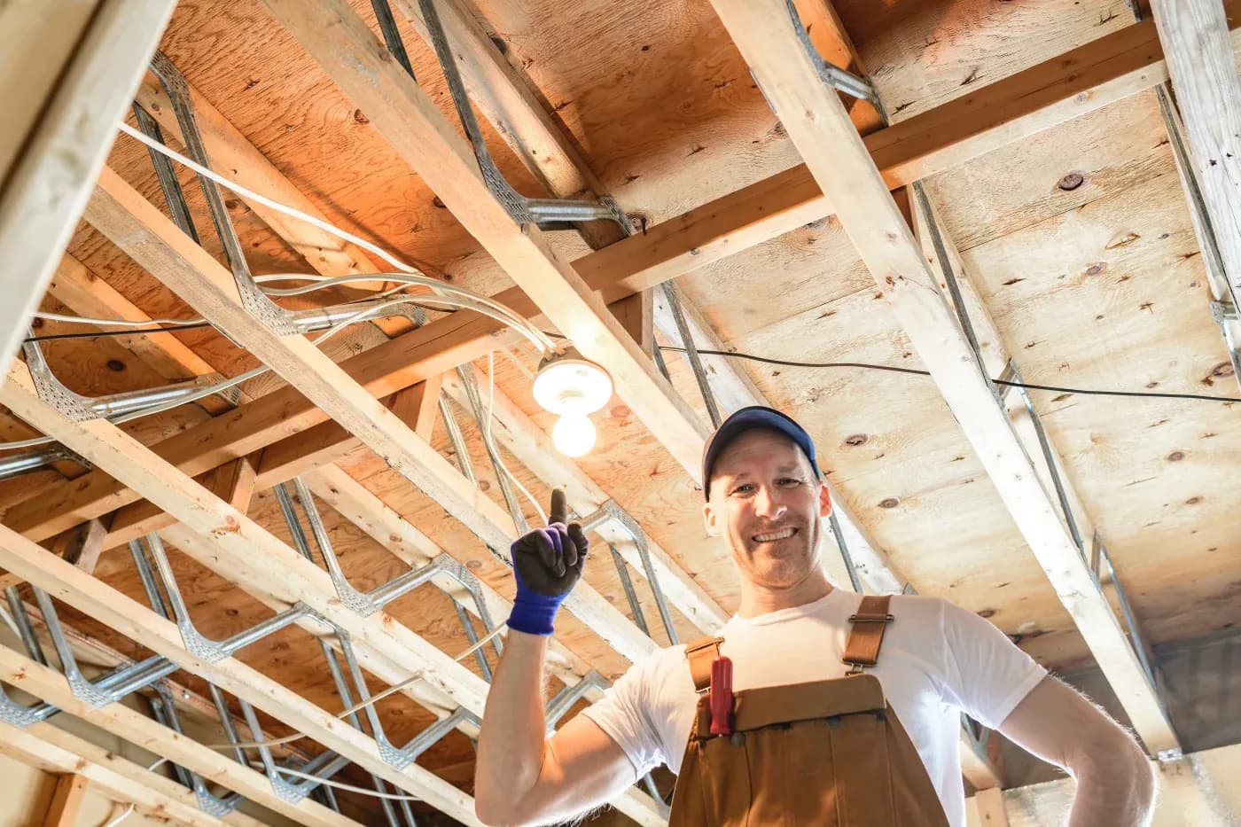 Smiling worker in overalls, wearing a cap, points at a lit bulb in a wooden ceiling with exposed wiring. Bright lighting, cheerful mood.