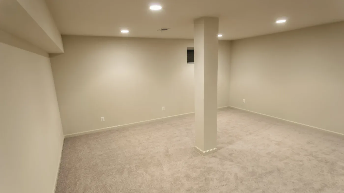 An empty, beige-carpeted basement with cream-colored walls, recessed ceiling lights, and a lone central support column conveys a neutral, unfinished tone.