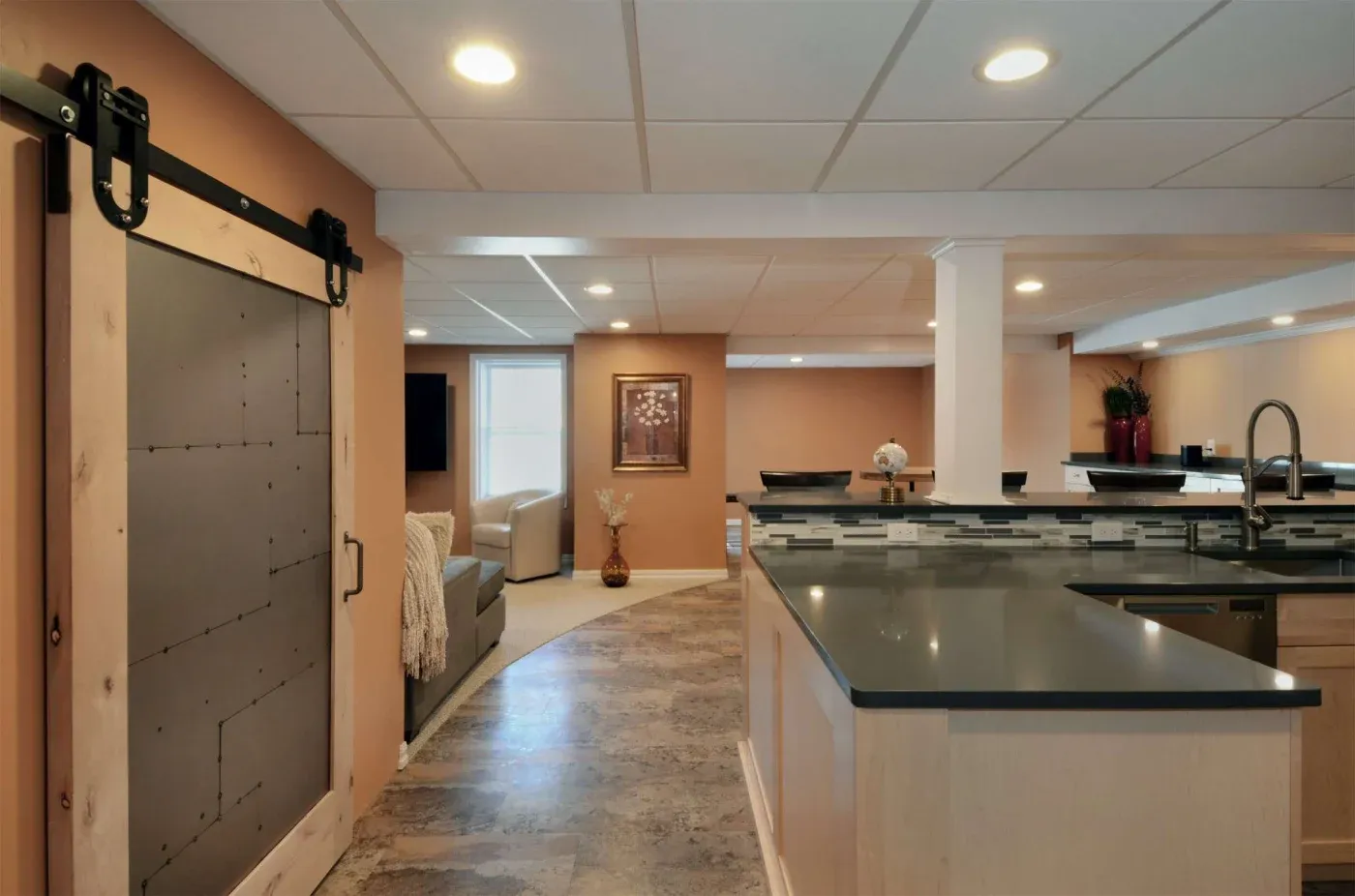 Modern basement with elegant decor. Features a sliding barn door, tiled floors, a sleek kitchen island, and a cozy seating area with soft lighting.