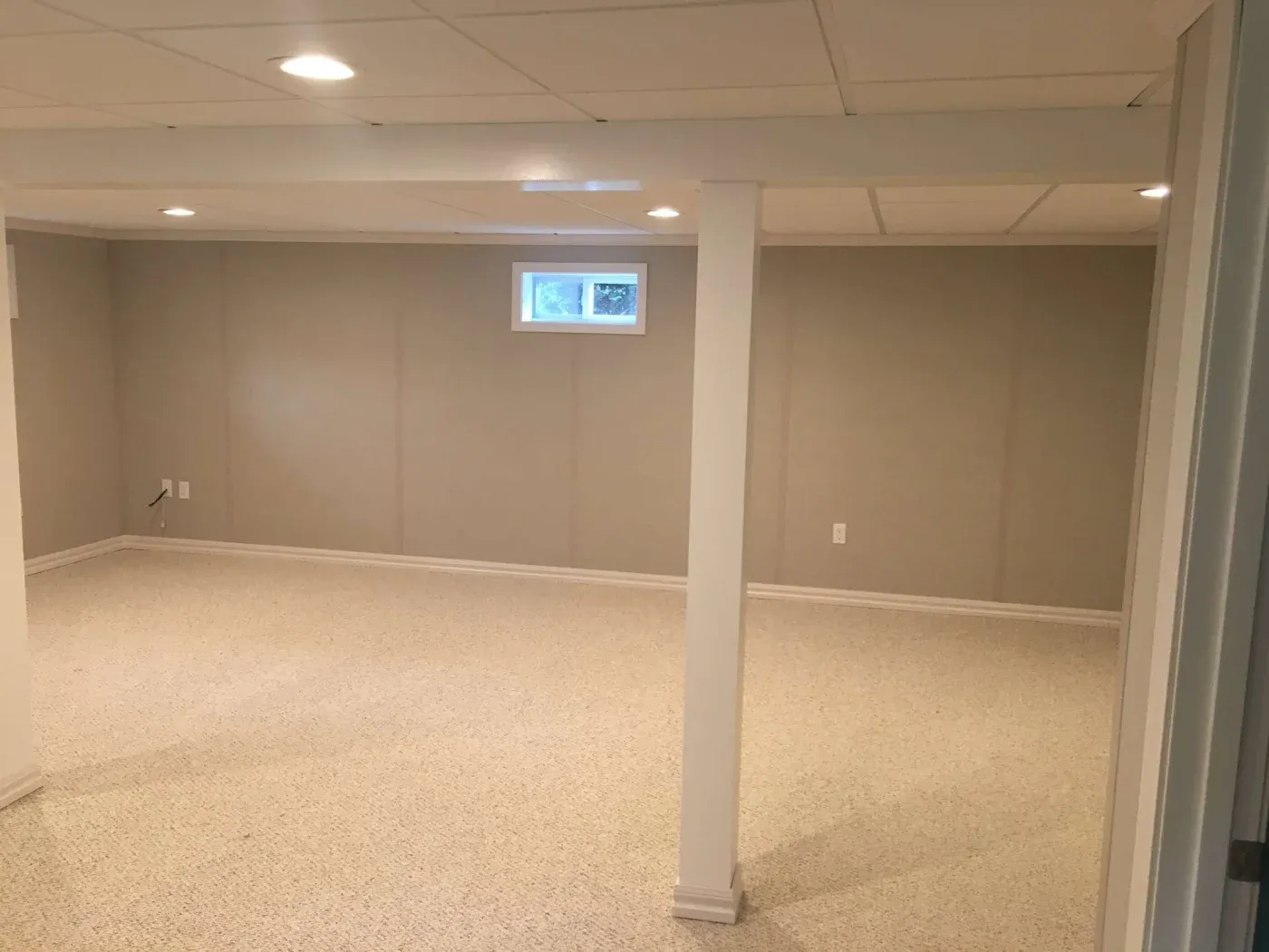 Finished basement with carpeted floor, beige walls, recessed ceiling lights, support column, and small window near ceiling