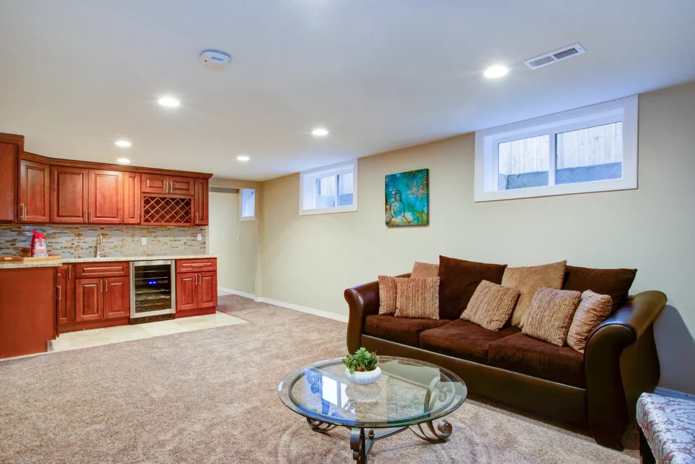 Cozy basement room with brown sofa and pillows, glass coffee table, and warm lighting. Modern kitchenette with wine rack and earthy decor.