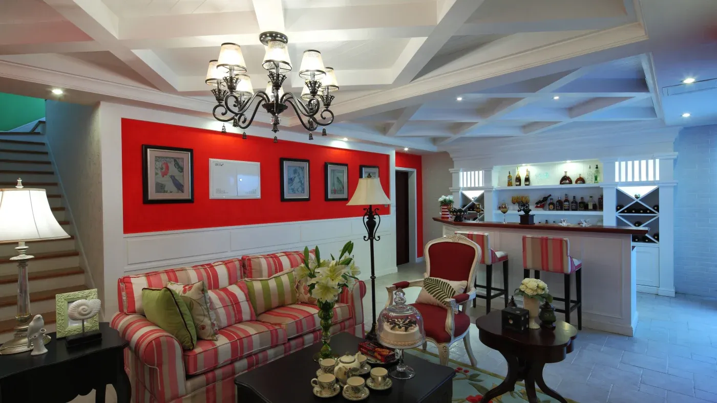 A cozy living room with a striped sofa, red and green cushions, and a flower centerpiece. A bar with bottles is in the background. Chandeliers hang from a coffered ceiling, creating an inviting atmosphere.