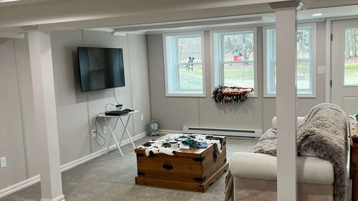 Bright, cozy living room with a wall-mounted TV, large windows, floral wreath, cream sofa, and vintage wooden trunk coffee table.