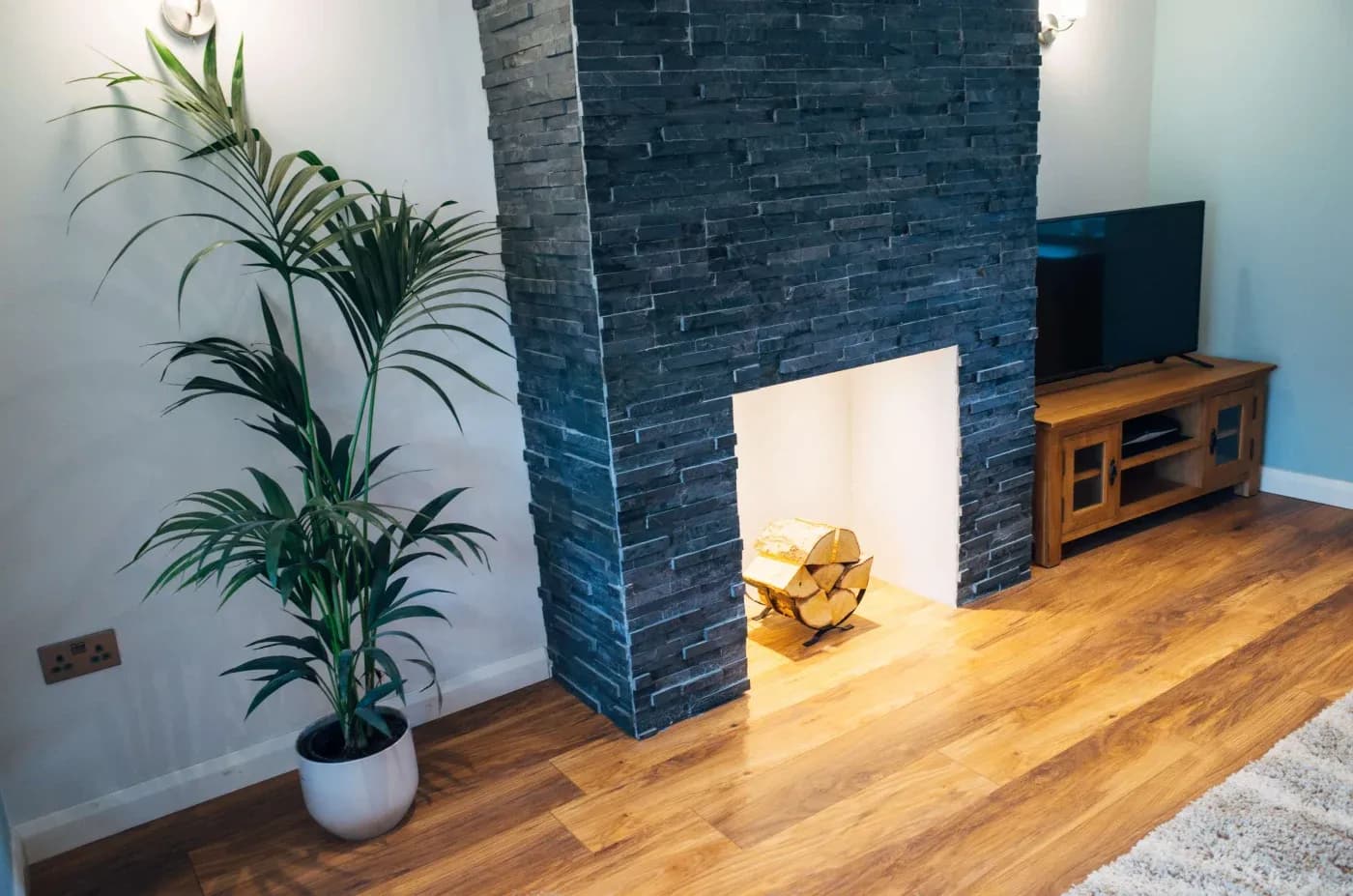 Modern living room with a dark stone fireplace, a potted palm, and a wooden TV cabinet. Warm wood flooring and soft lighting create a cozy ambiance.