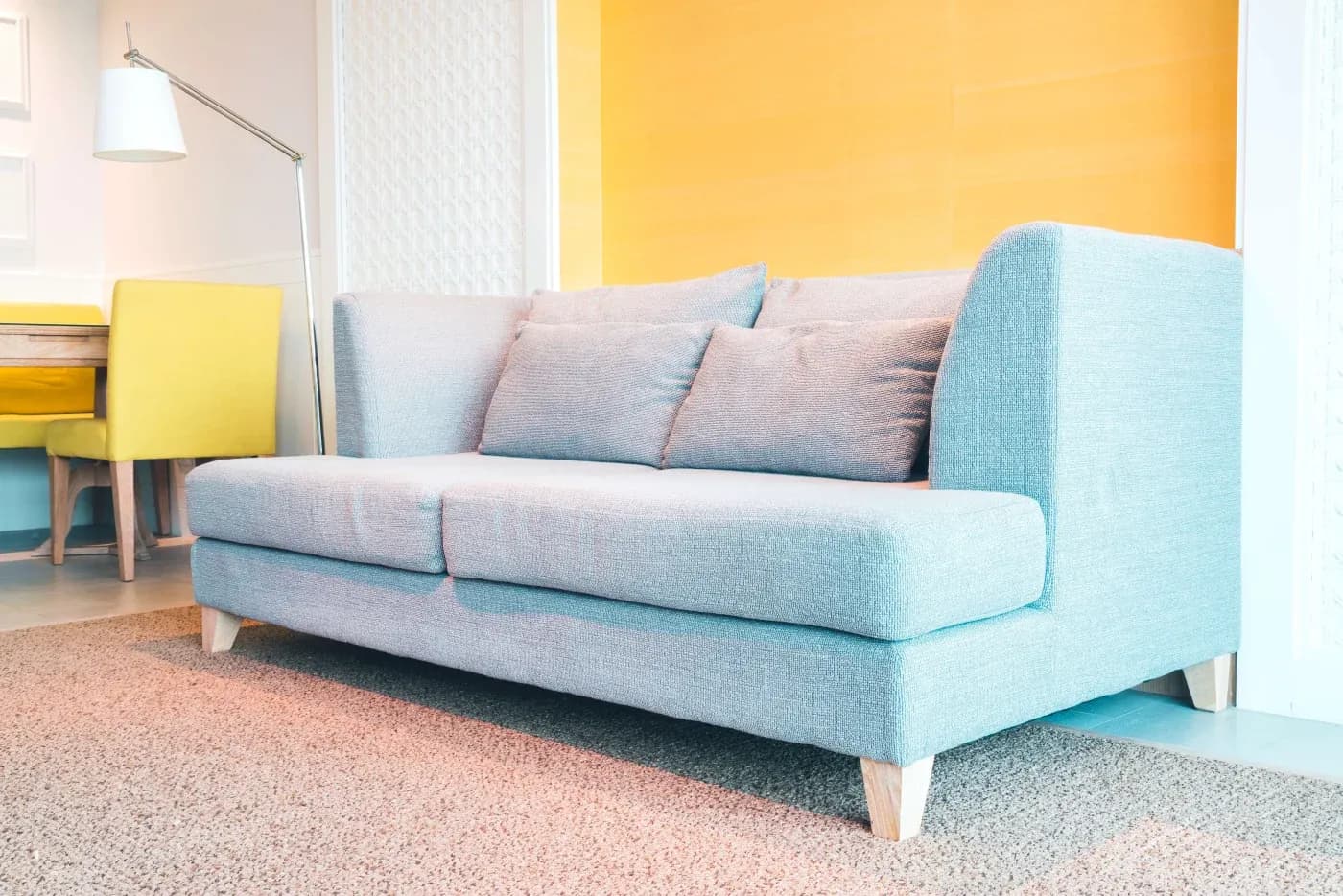 Modern living room with a light blue sofa, wooden legs, and three cushions. A yellow wall and armchair add brightness, creating a cozy, inviting atmosphere.