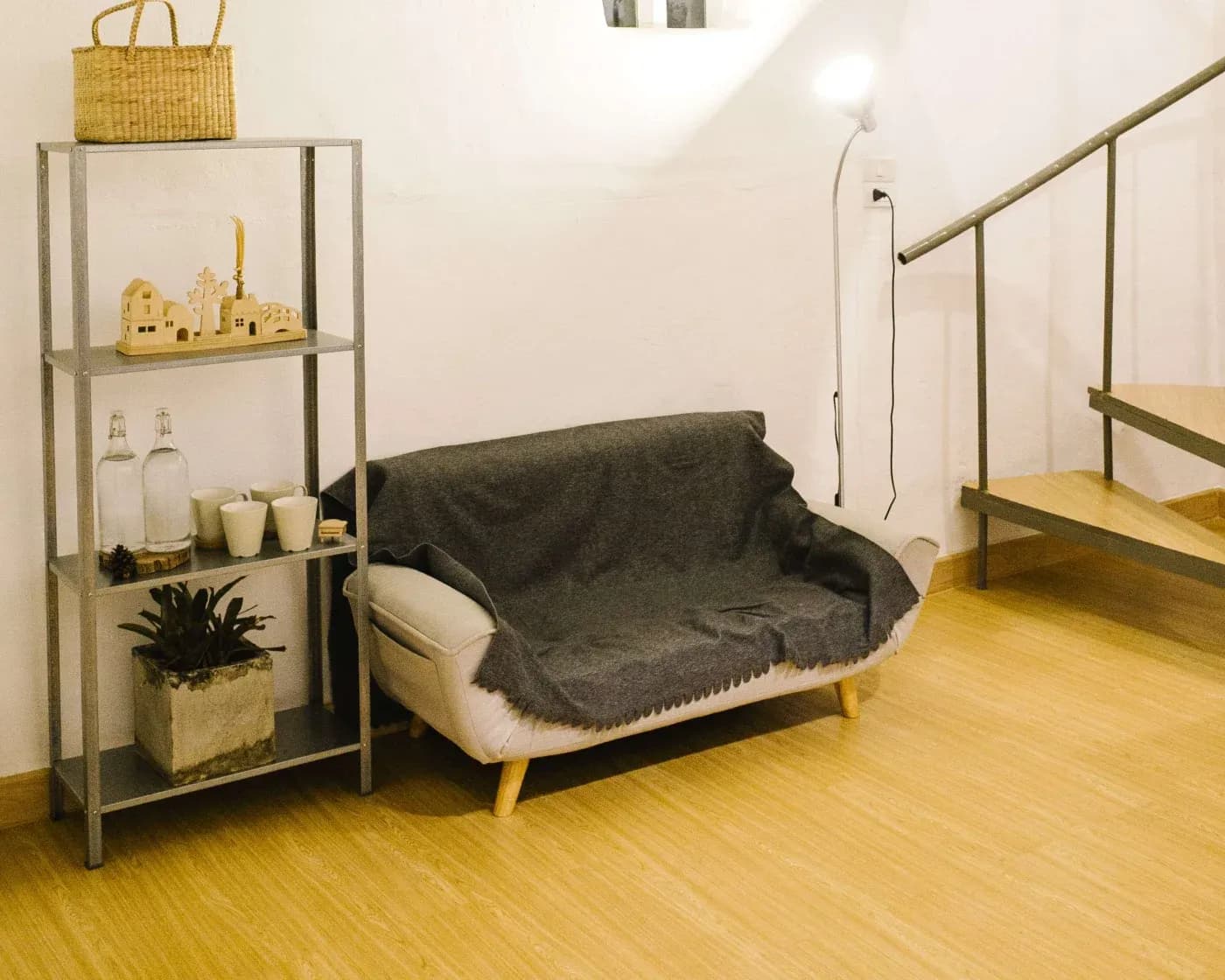 Minimalist living room corner with a small gray couch, wooden shelf holding decor and plants, a floor lamp, and wooden stairs with metal railing.