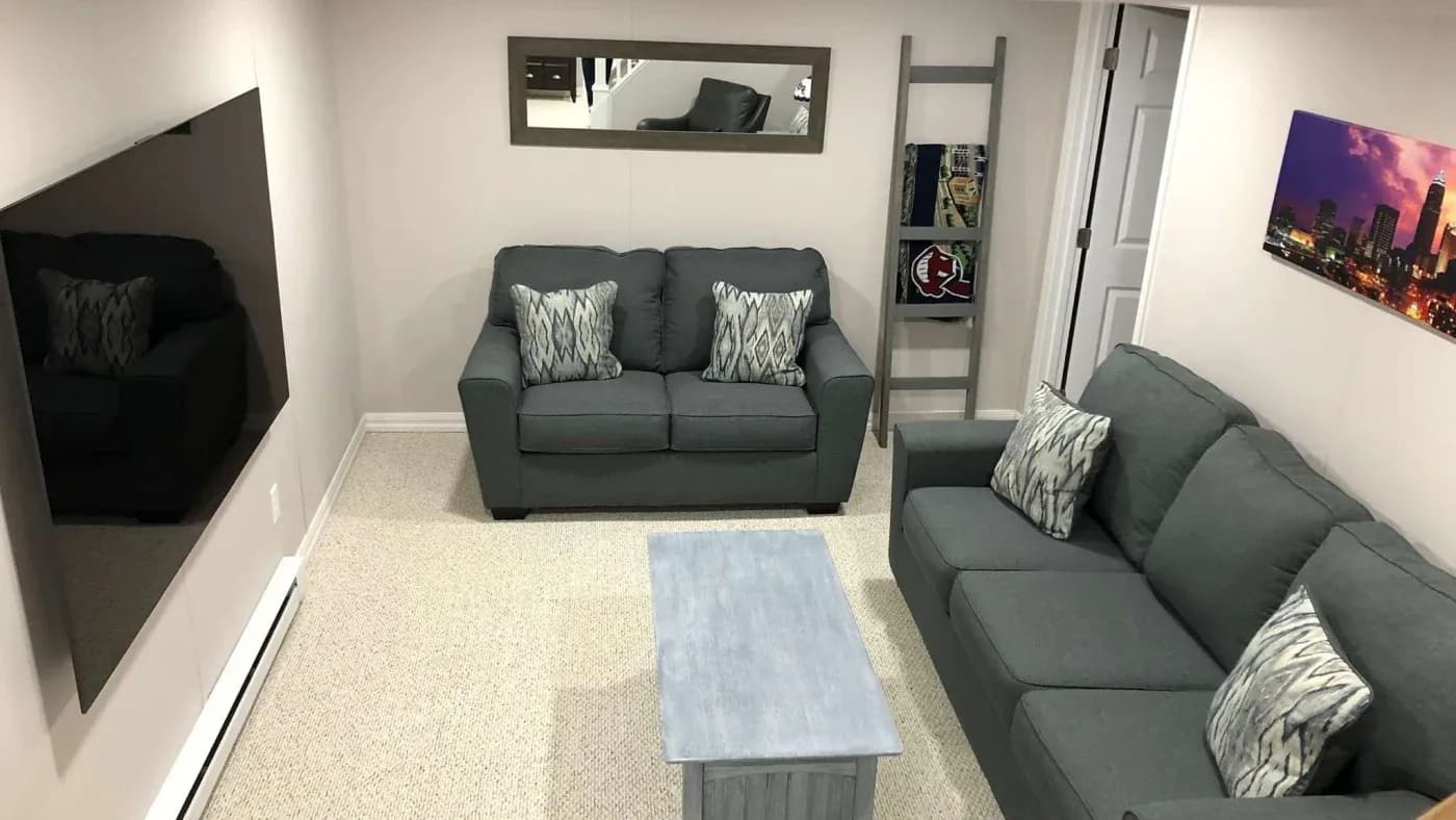 A cozy living room with gray sofas and patterned pillows, a light blue coffee table, wall-mounted TV, and cityscape artwork. The ambiance is calm and inviting.