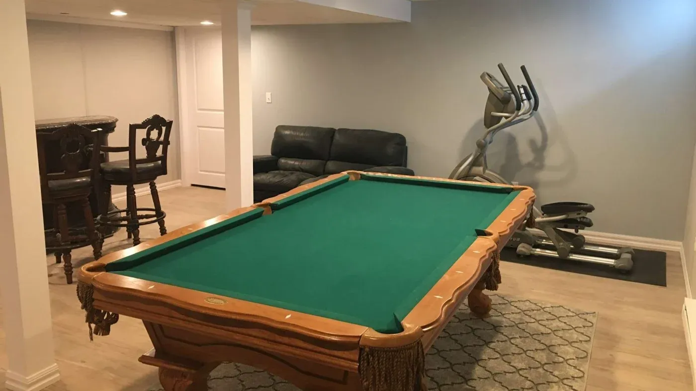 A cozy basement with a pool table at the center, a black leather couch by the wall, an elliptical machine, and two bar stools with a small table.