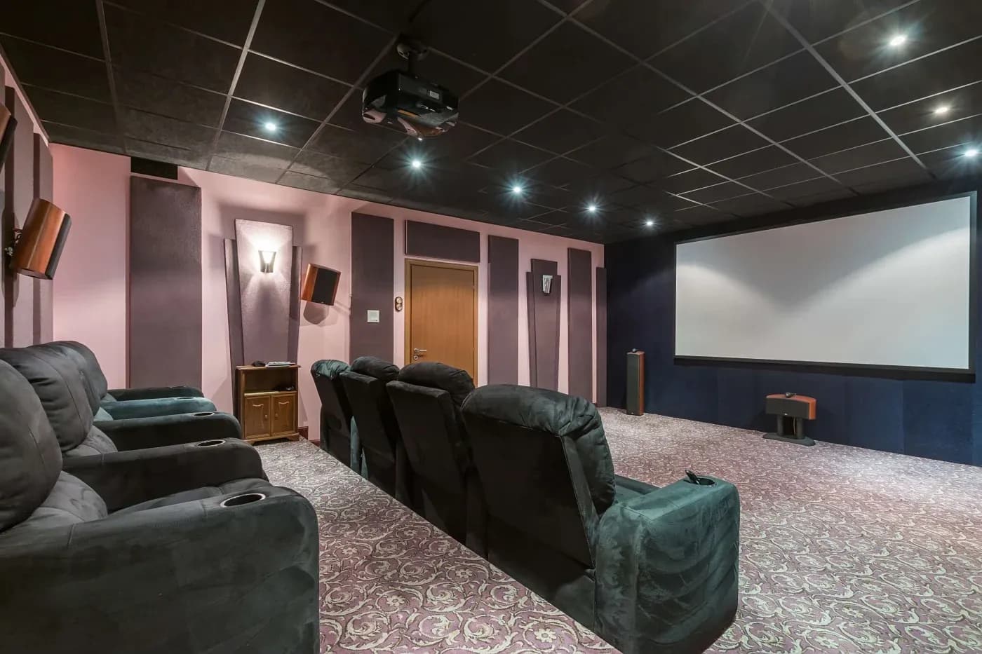 A plush home theater with a row of black recliner seats, soft pink and black walls, and a large projector screen, creating a cozy, inviting atmosphere.