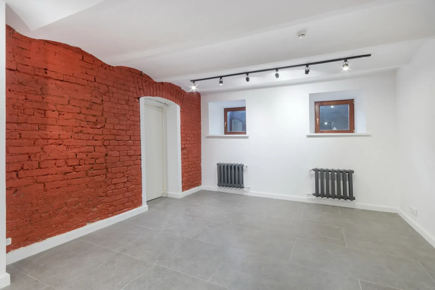 Minimalist room with red brick accent wall, gray tile flooring, and white walls. Two small windows and radiators, with track lighting above.