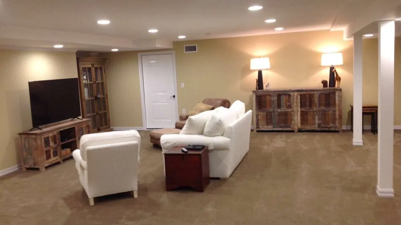 A cozy basement living room with beige walls, a white sofa, and an armchair. A large TV sits on a wooden stand, and lamps illuminate the space warmly.
