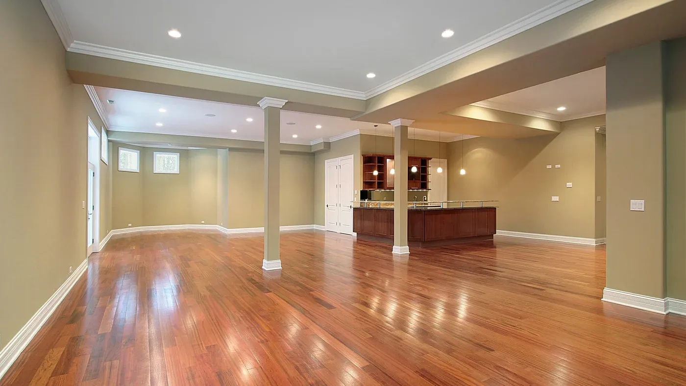 Spacious open-concept basement with polished hardwood floors, beige walls, and recessed ceiling lights. A modern kitchen area is seen in the background.