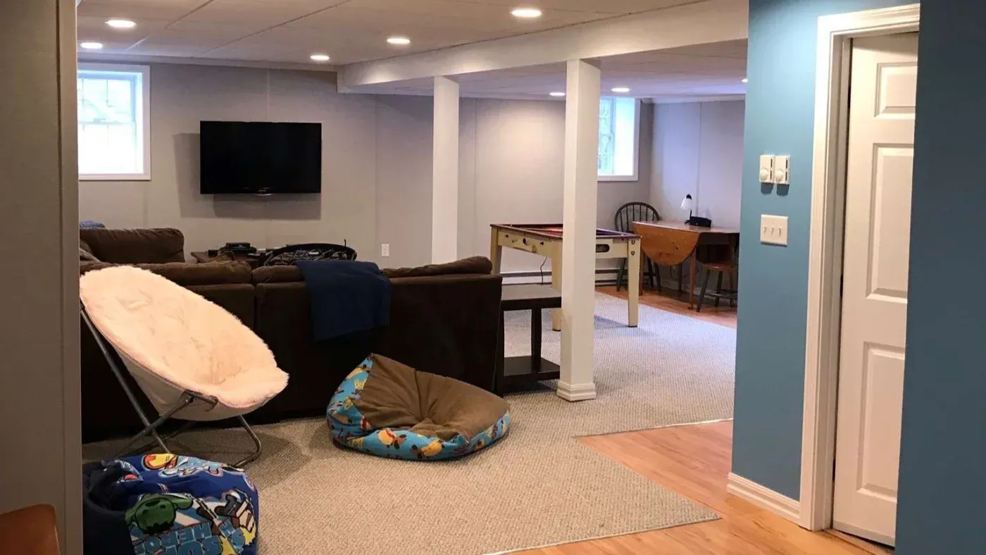 A cozy basement with a sectional sofa facing a wall-mounted TV. A game area with a table and chairs is visible. Relaxed, inviting atmosphere.