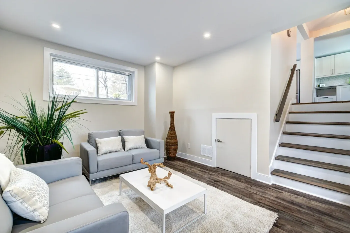 Modern living room with gray sofas, white pillows, and a sleek coffee table on a plush rug. A large plant and decorative vase add style. Stairs beside a small door lead to a bright kitchen, conveying a cozy and contemporary atmosphere.
