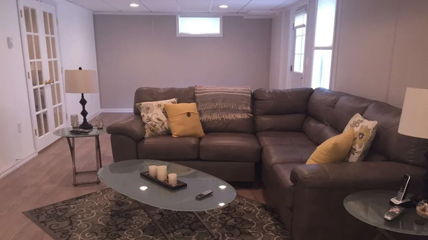 A cozy living room with a brown sectional sofa adorned with yellow and patterned pillows. Oval glass coffee table holds candles and remote. Soft, ambient lighting.