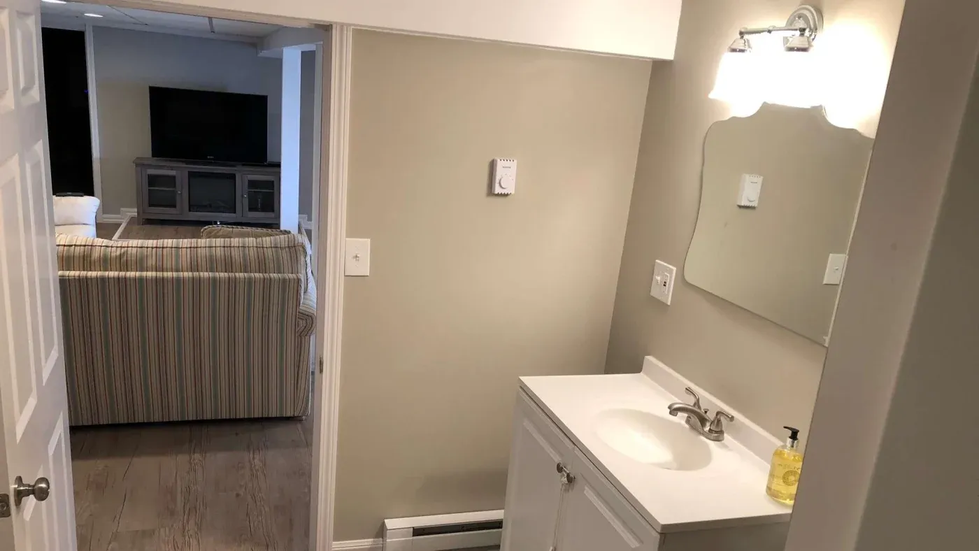 Small bathroom with white vanity, mirror, and wall lamp. Adjacent room features a striped sofa and TV. Neutral tones create a calm, cozy atmosphere.