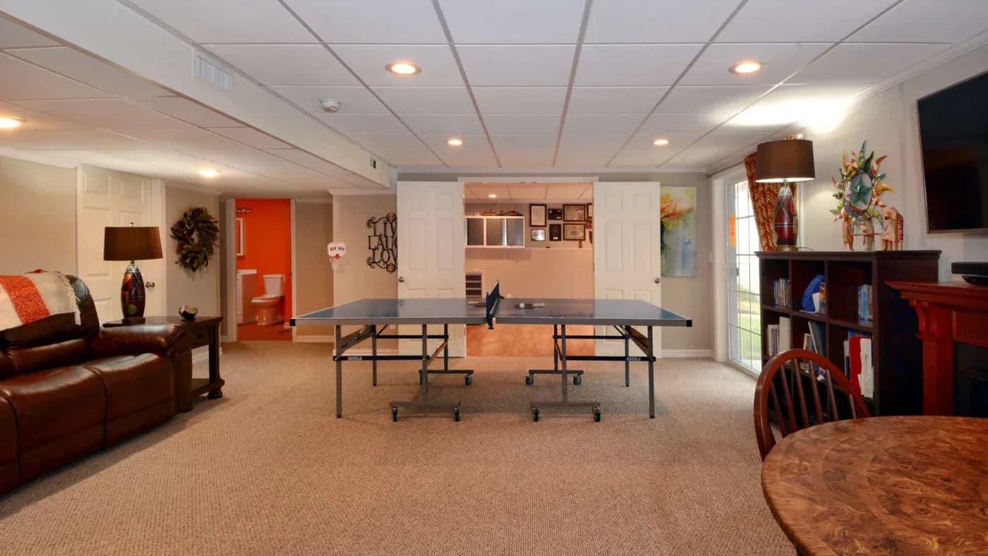 Basement with ping pong table, brown leather sofa, and round wooden table. Warm lighting creates a cozy, recreational atmosphere.