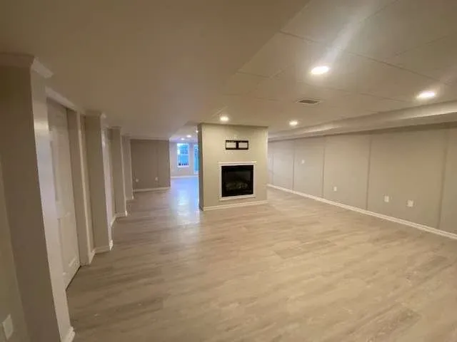 Spacious, well-lit basement with light wood flooring, recessed lighting, and a centered modern fireplace. The tone is clean and inviting.