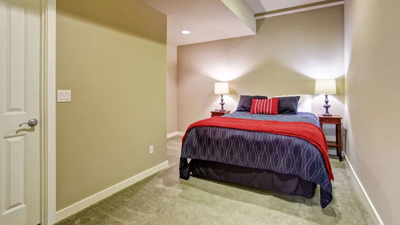 Cozy bedroom with beige walls and carpet. A neatly made bed with a red blanket and pillows sits against the wall, flanked by wooden nightstands with lamps.
