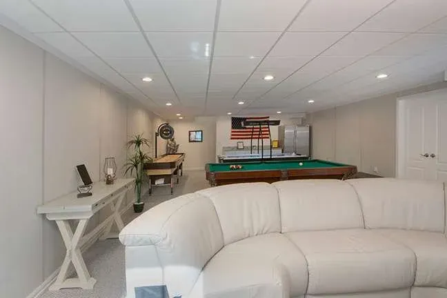A cozy basement game room with a white curved sofa, pool table, and foosball table. American flag wall decor; bright with recessed lighting.