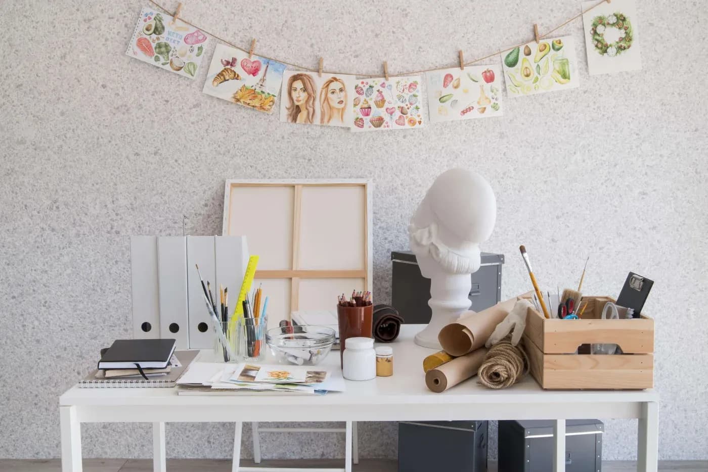 Art studio with a white table, art supplies, and a bust sculpture. Watercolor paintings hang above, creating a creative and inspiring atmosphere.