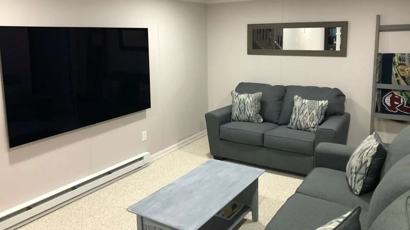 A cozy living room with a large, wall-mounted TV, two gray sofas, a light blue coffee table, and a mirror above one sofa. The atmosphere is calm and inviting.