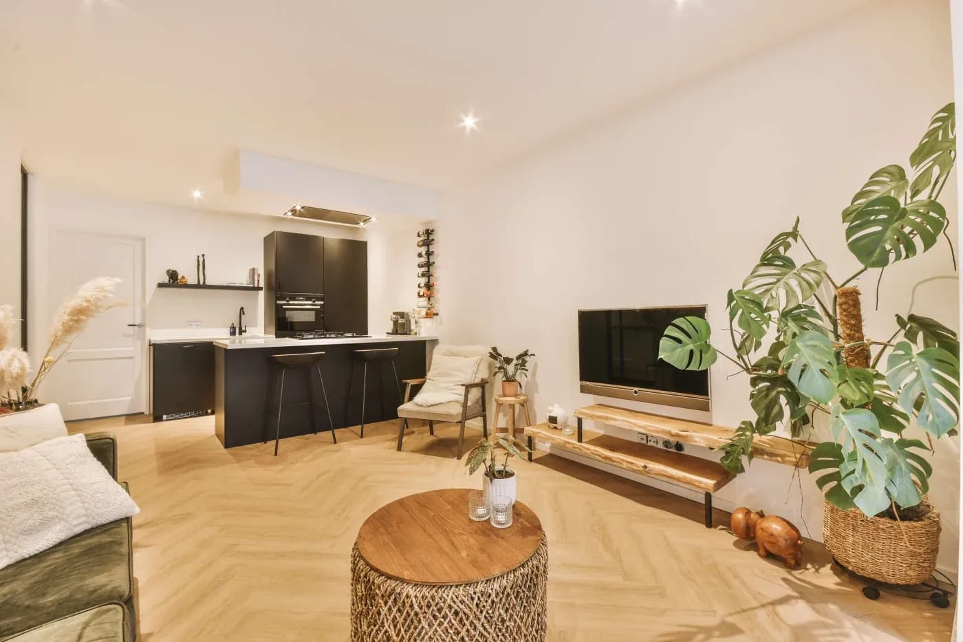 Modern living room with beige walls, herringbone wooden floors, a black kitchen area, a TV on a wooden stand, a leafy plant, and cozy seating.