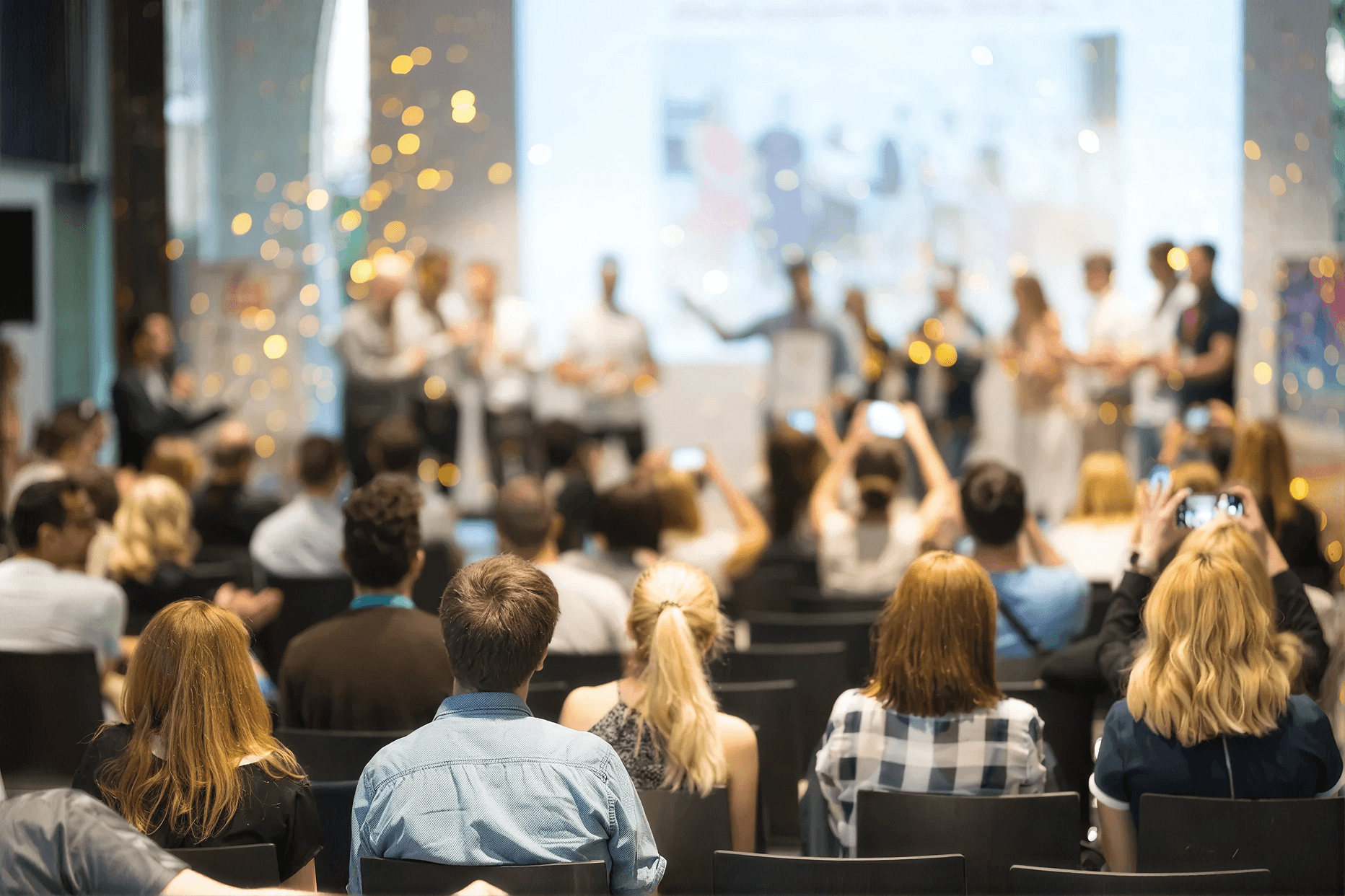 Audience seated at a conference or award ceremony, with blurred figures on stage. Attendees capture moments on phones. Atmosphere is lively and celebratory.