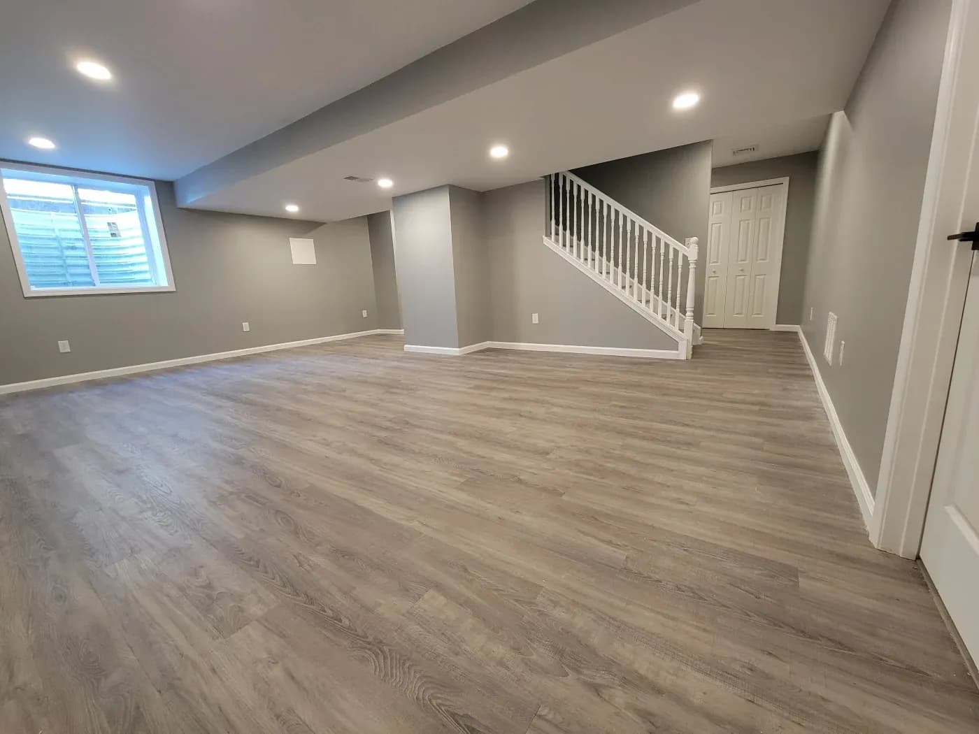 Spacious, modern basement with gray walls, wood flooring, and recessed lighting. A staircase with white railings leads upstairs. A window offers natural light.
