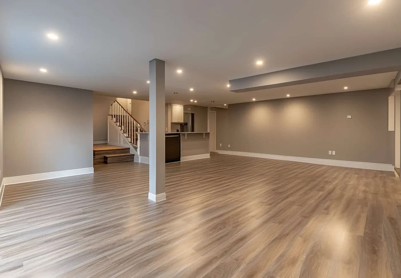 Spacious empty basement with wood flooring and recessed lighting. Features a central column, neutral walls, and a staircase leading upstairs. Modern and clean design.