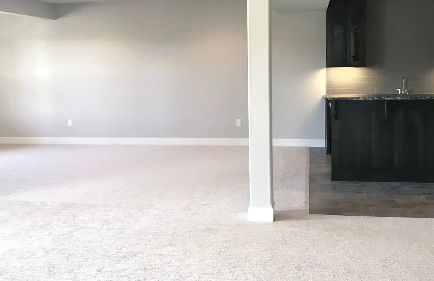 A spacious, empty room with light gray walls and beige carpet, featuring a white beam. On the right, a small kitchen area with dark cabinets and a tiled floor.